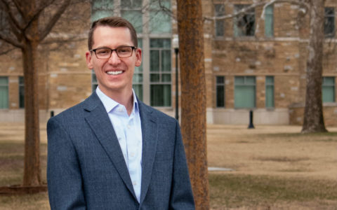 a man standing outdoors wearing glasses, a light blue shirt and a dark blue blazer