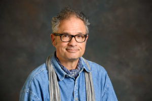 Man smiling and wearing glasses while wearing an open-collar blue shirt and a neck scarf