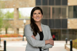 young woman with long dark hair wearing a grey pin-stripe pant suit standing with arms crossed while smiling