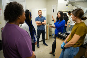 A small group of people standing in a room listening to a woman in a blue shirt speak