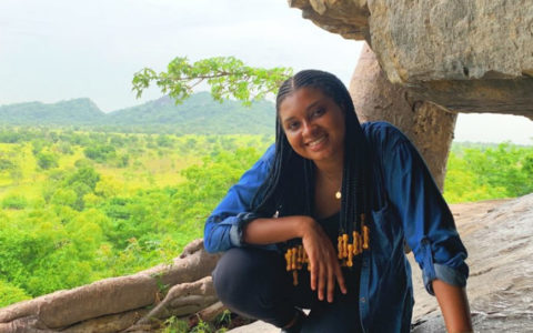 woman with long black hair crouching near a rock formation