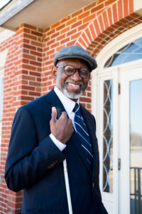 man standing in front of a brick building wearing a grey cap, white shirt, striped tie and blue blazer