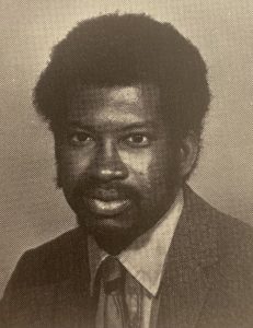 Black and white photo of a young man with dark hair wearing a light-colored shirt and dark tie and blazer