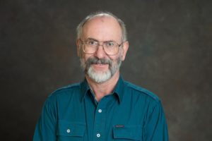 man with short grey hair and beard wearing glasses and a teal blue shirt