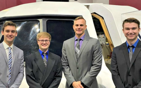 four young men in suits and ties standing in front of a space shuttle cockpit