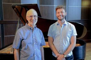 older gentleman in a blue shirt standing beside a younger man in a blue shirt in front of a grand piano