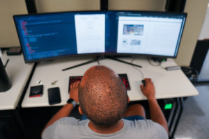 photo from above and behind of a young man seated at a desk in front of two computer monitors