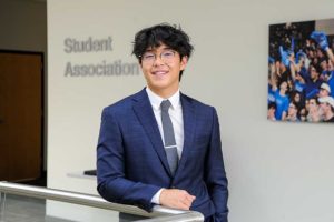 young man with black hair wearing glasses, white shirt, grey tie, and a blue blazer