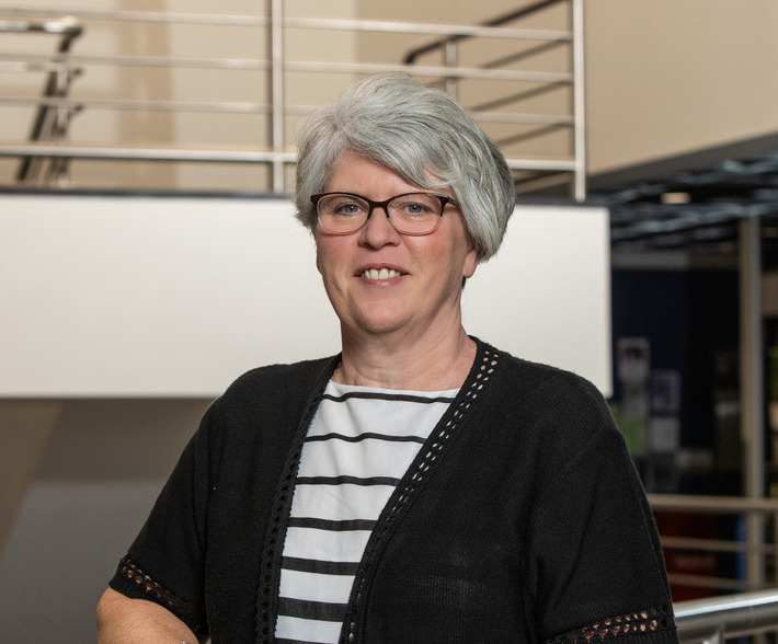 Photo of Kimberly Adams smiling with brown glasses and white hair inside of Keplinger Hall.