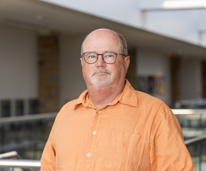 Photo of Greg Gardner with gray glasses with orange shirt inside of Student Union.