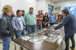 A dozen men gathered around a wooden table looking at black-and-white photographs