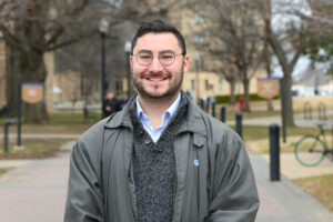 young man with short hair and a trim beard and round-frame glasses wearing a grey jacket while standing outdoors