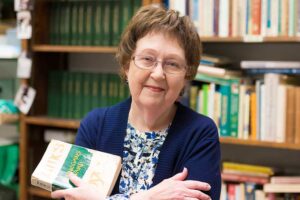 woman with short hair and glasses wearing a blue top and holding a book entitled Finnegan's Wake