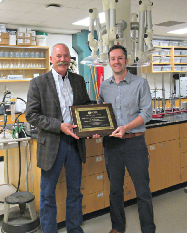 two men standing in a chemistry lab holding an award plaque