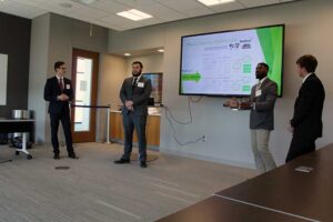 four young men standing several feet apart from each other in a room in front of a large screen on which is projected a PowerPoint slide