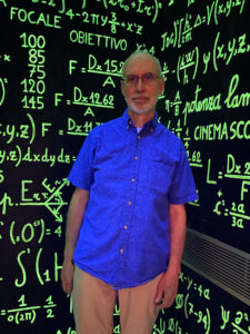 man in a blue short-sleeved shirt standing against a black background emblazoned with bright green mathematical formulae