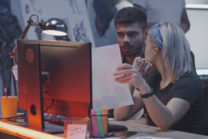 two young people at a desk discussing images drawn on paper and a computer monitor