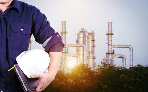 partial view of a man holding a white hardhat in front of an oil refinery with the sun shining through it