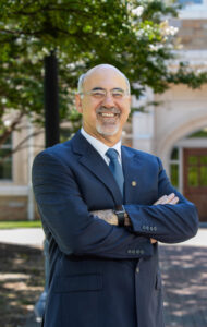 Dean in a suit stands with arms crossed in front of a building.