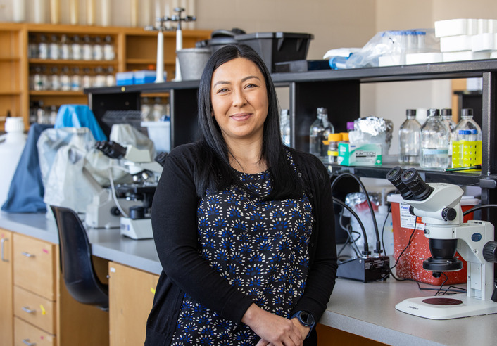 Biological science researcher stands in a lab with microscopes. DoD award recipient.