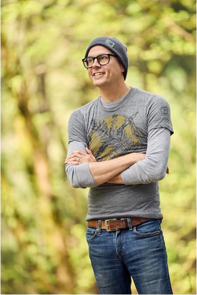 Author in a gray beanie and glasses stands outdoors with arms crossed, smiling.