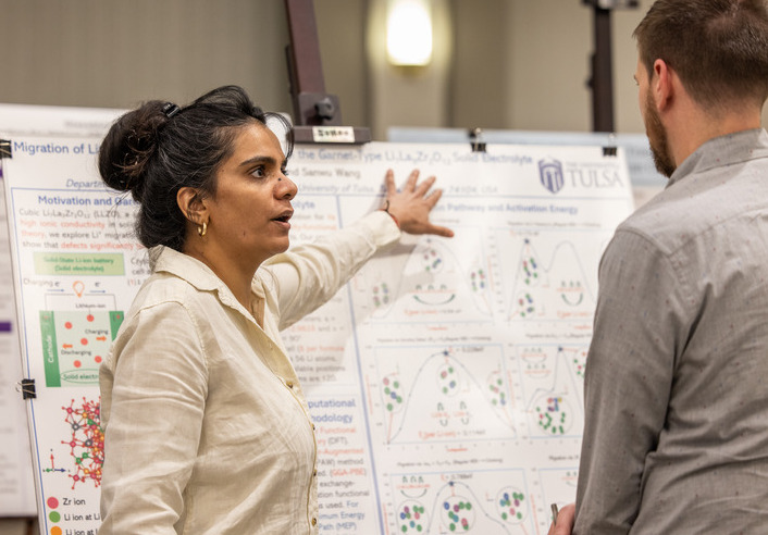 Physics doctoral student presenting battery research at UTulsa, explaining charts on a poster board.