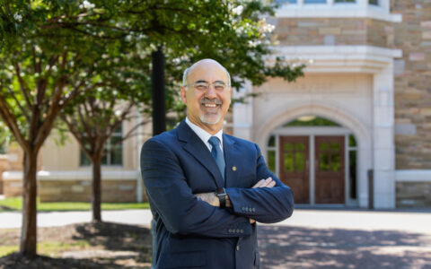 University president in suit, arms crossed, standing in front of building.