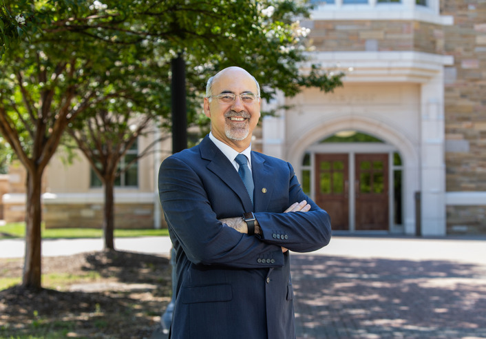 University president in suit, arms crossed, standing in front of building.