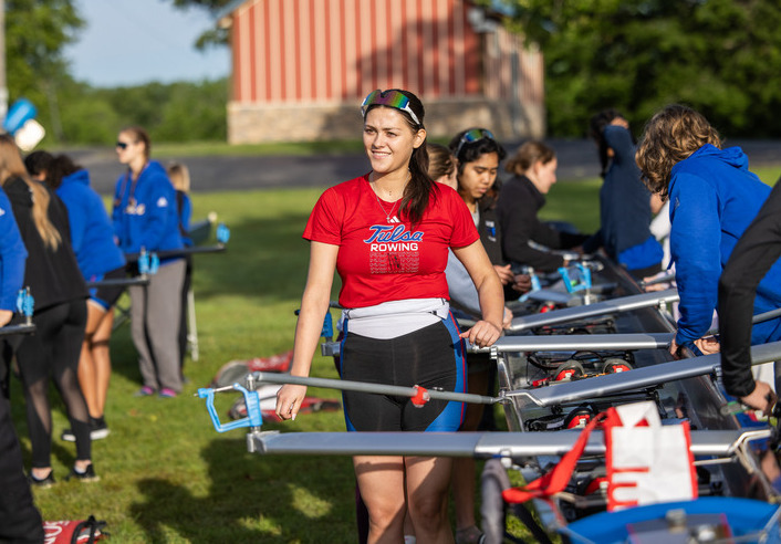 Tulsa Rowing team member smiles, preparing equipment for practice. "Tulsa Rowing" text on shirt.