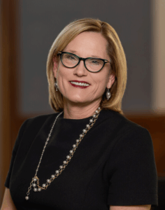 Professional portrait of a woman with blonde hair, wearing glasses and a pearl necklace. 