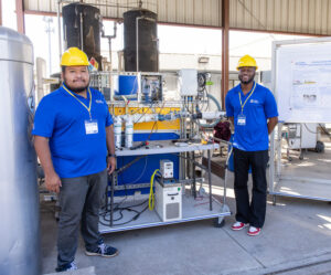 Two Cyber Fellows Community members stand by their project, wearing hard hats and blue shirts.