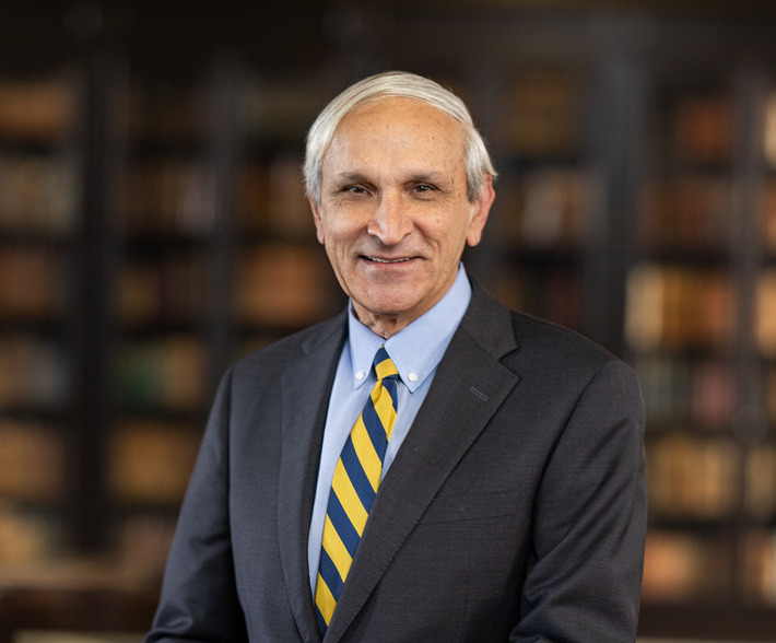 Portrait of Ken McQueen wearing a suit and tie, standing in front of a bookshelf.