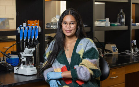 UTulsa NSF research: Woman in lab coat and gloves sits beside a microscope.