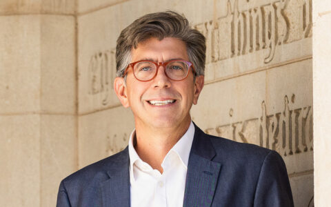 Dean of Arts & Sciences Blaine Greteman smiles in front of stone wall with carved letters.