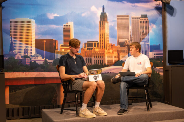 Two TUTV students sitting with laptops, Tulsa skyline backdrop.