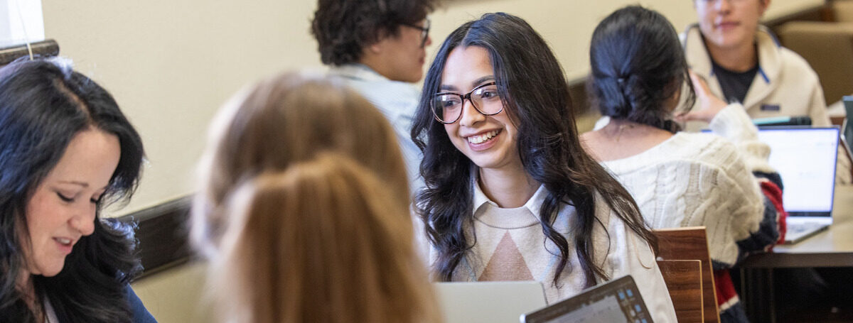 Smiling graduate student in glasses studying with classmates at a table.