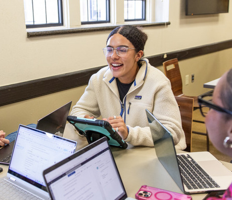 Smiling student with glasses at a table with laptops. Collaborative graduate admission work.