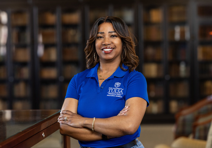 OCII staff member in a blue shirt with the University of Tulsa logo, standing in front of bookshelves.