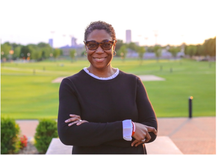 Smiling woman in glasses with arms crossed in a park setting.