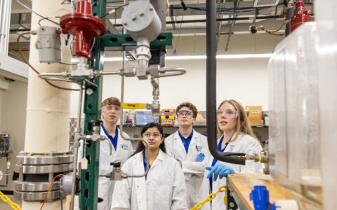 Engineering students in lab coats and safety glasses examine equipment in a College of Engineering lab.