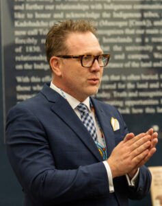 Dean wearing glasses and a suit, speaking with hands clasped, in front of a wall of text