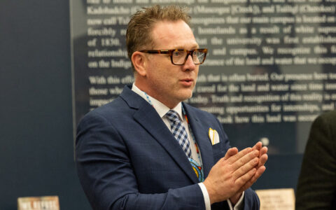 Dean speaks with hands clasped, wearing a suit and glasses.