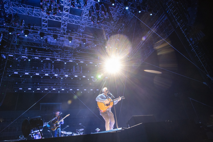Zach Bryan performs live in concert with guitar under bright stage lights.