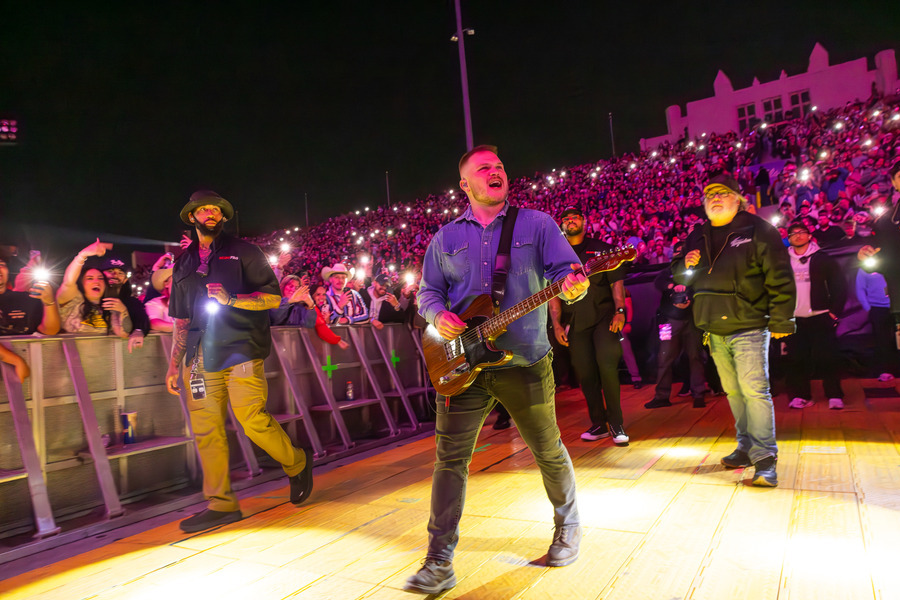 Zach Bryan performing live at a concert with a crowd holding up lights.