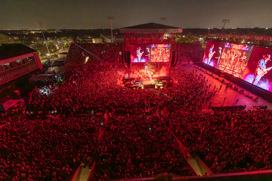 Aerial view of a packed Zach Bryan concert with red stage lights and large video screens.