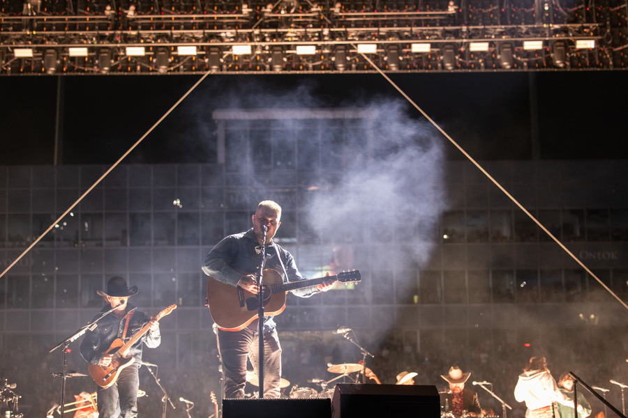 Zach Bryan performing live at a concert, illuminated by stage lights and smoke.