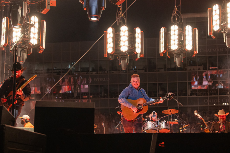 Zach Bryan performing live in concert with his band under stage lights.