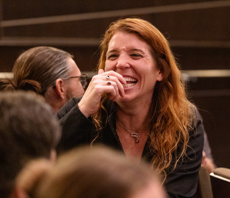 Woman laughing at an event, with red hair and a black jacket.