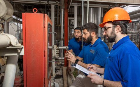 Three students inspect equipment and pipes; one is wearing a hard hat