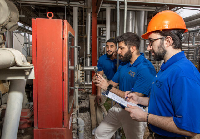 Three students inspect equipment and pipes; one is wearing a hard hat 